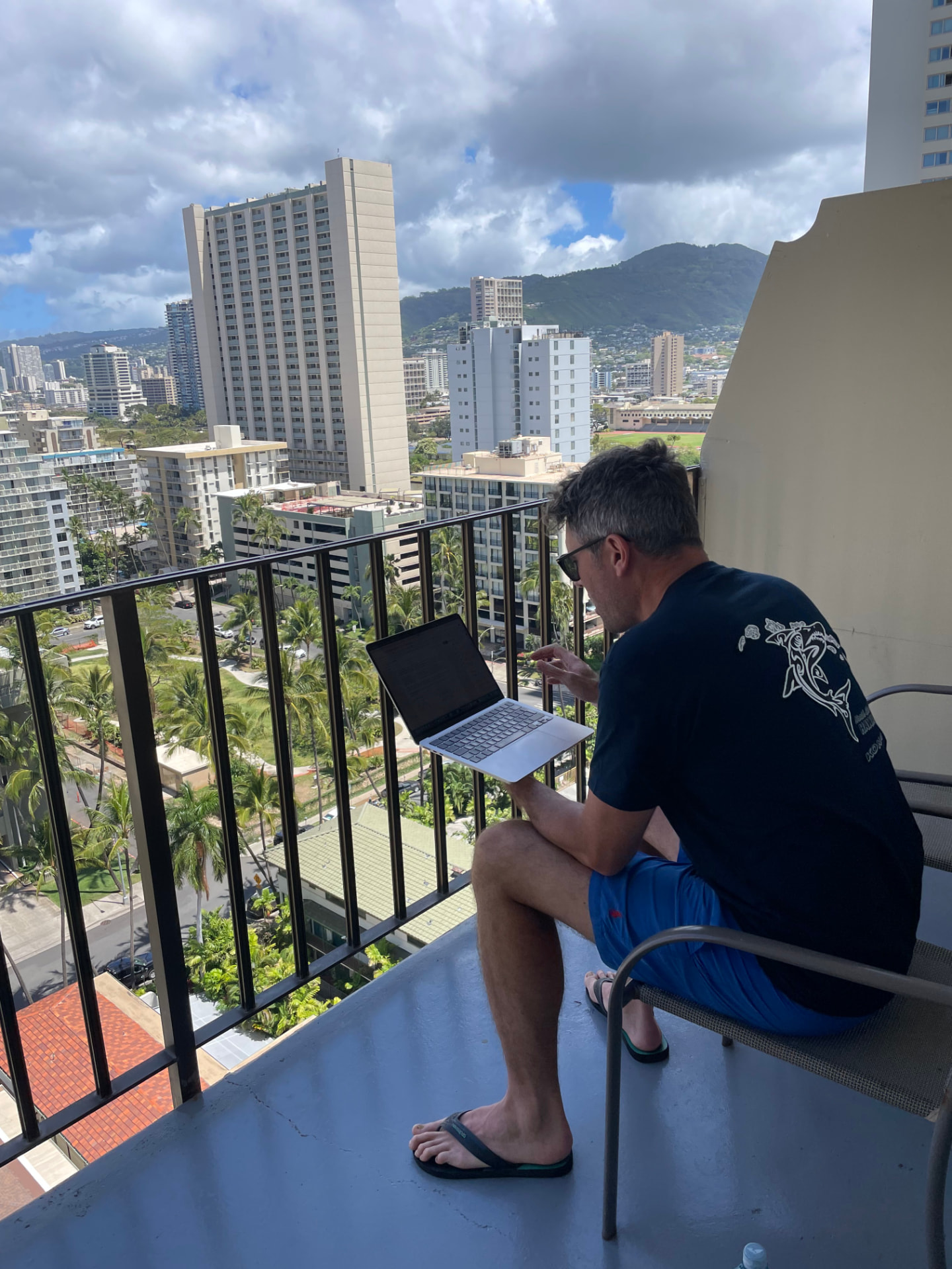 Nigel working on a laptop on a high balcony overlooking the Honolulu skyline and mountains.