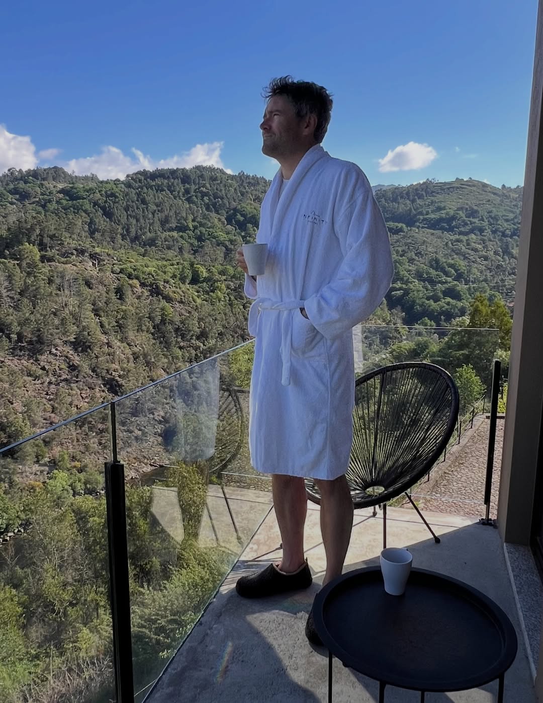 Nigel in a white robe holding a mug on a balcony overlooking wooded hills in Paradamonte, Portugal.