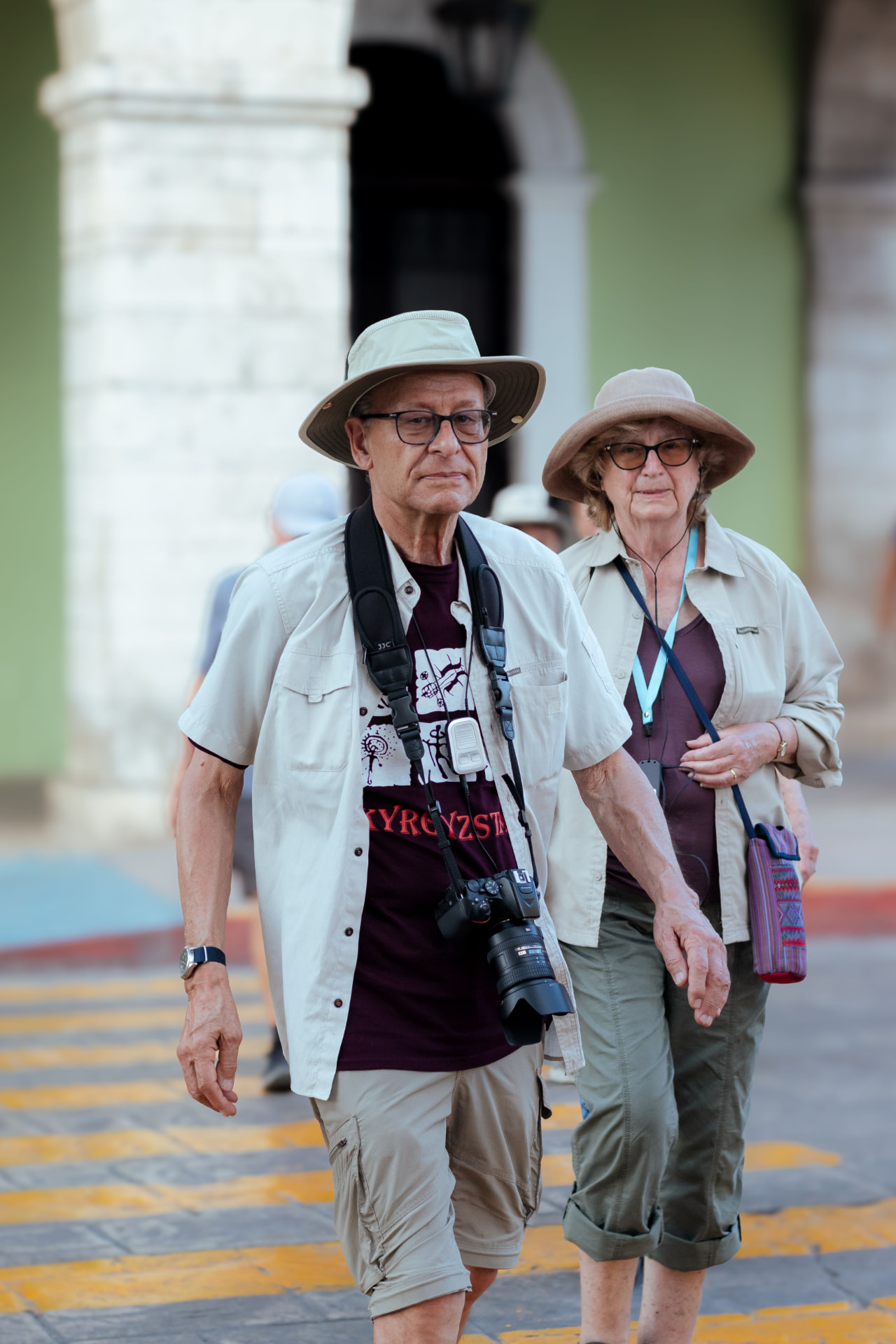 An older couple in sun hats walking across a colonial square with cameras around their necks.