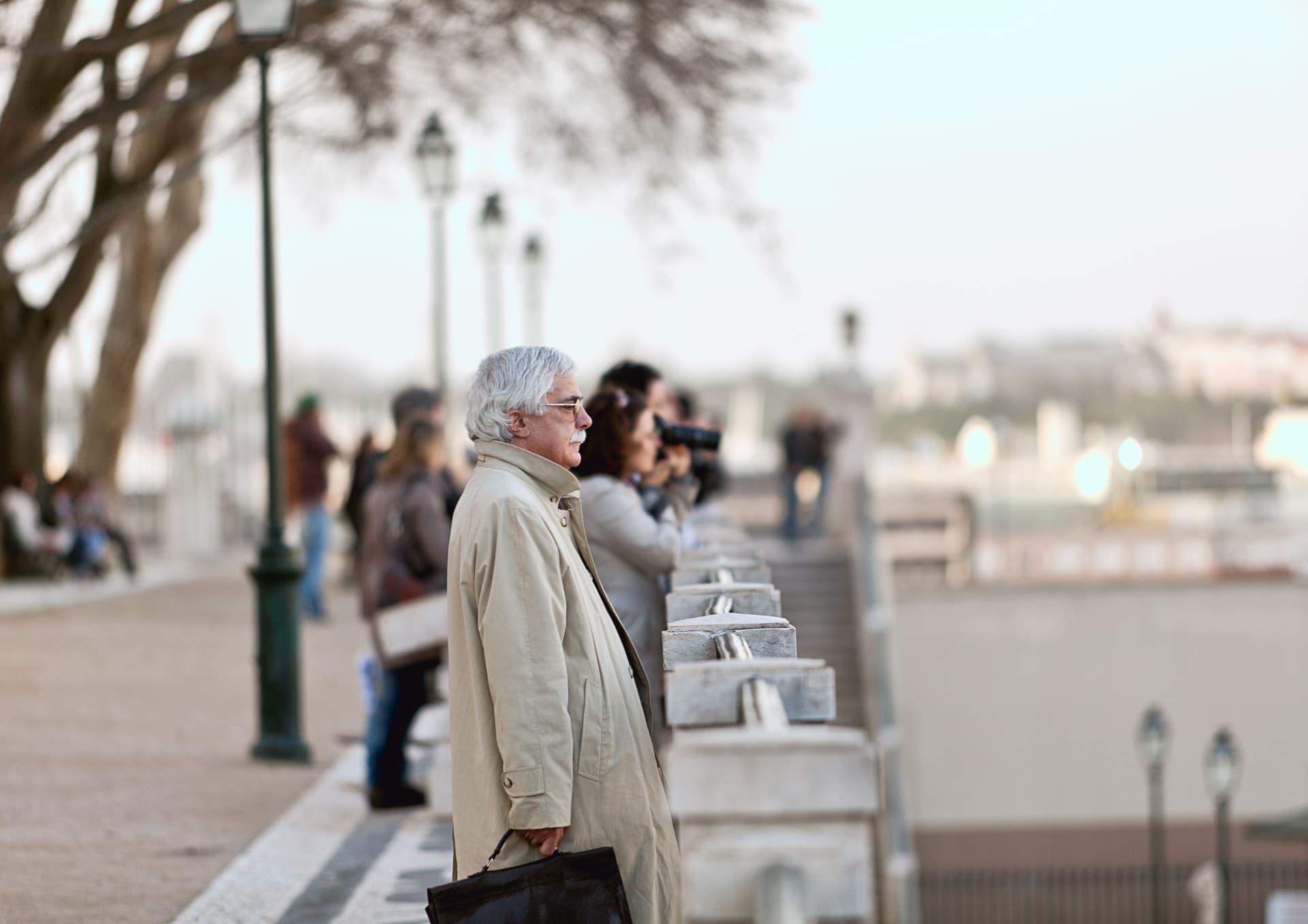 A man in a long coat standing at a riverside balustrade, watching the view.