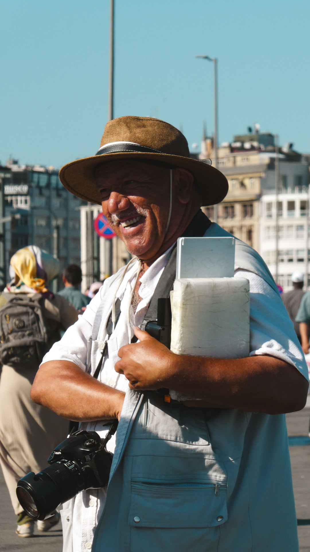 A smiling older man in a straw hat carrying a camera across a busy waterfront.