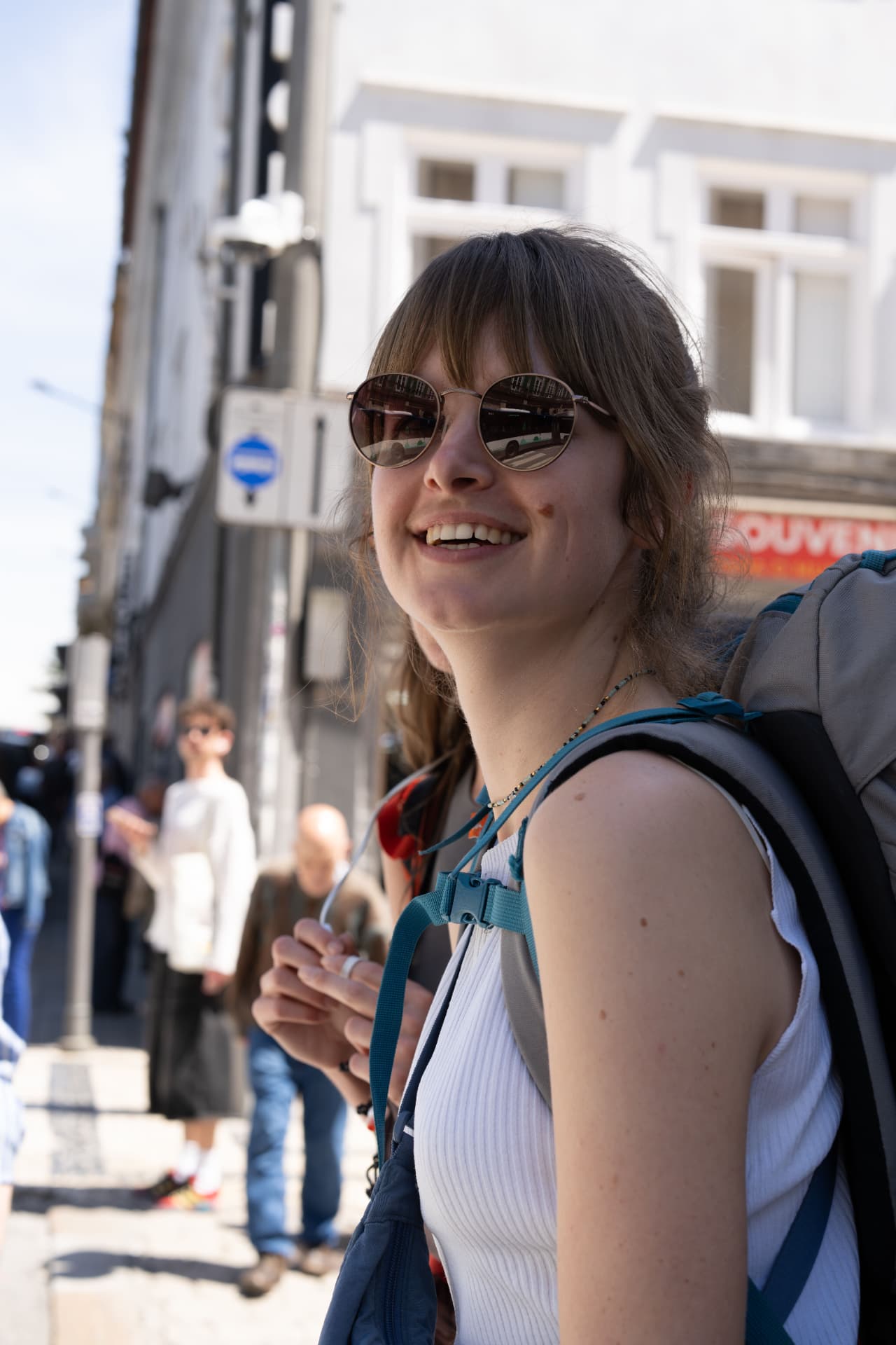 A traveller in sunglasses laughing on a sunny city street, backpack on her shoulders.