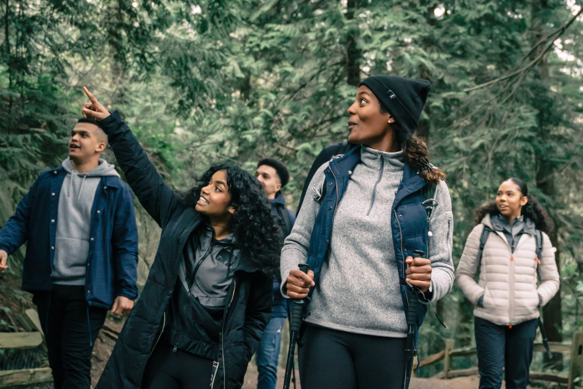 A group of friends hiking a forest trail, one pointing up through the trees.
