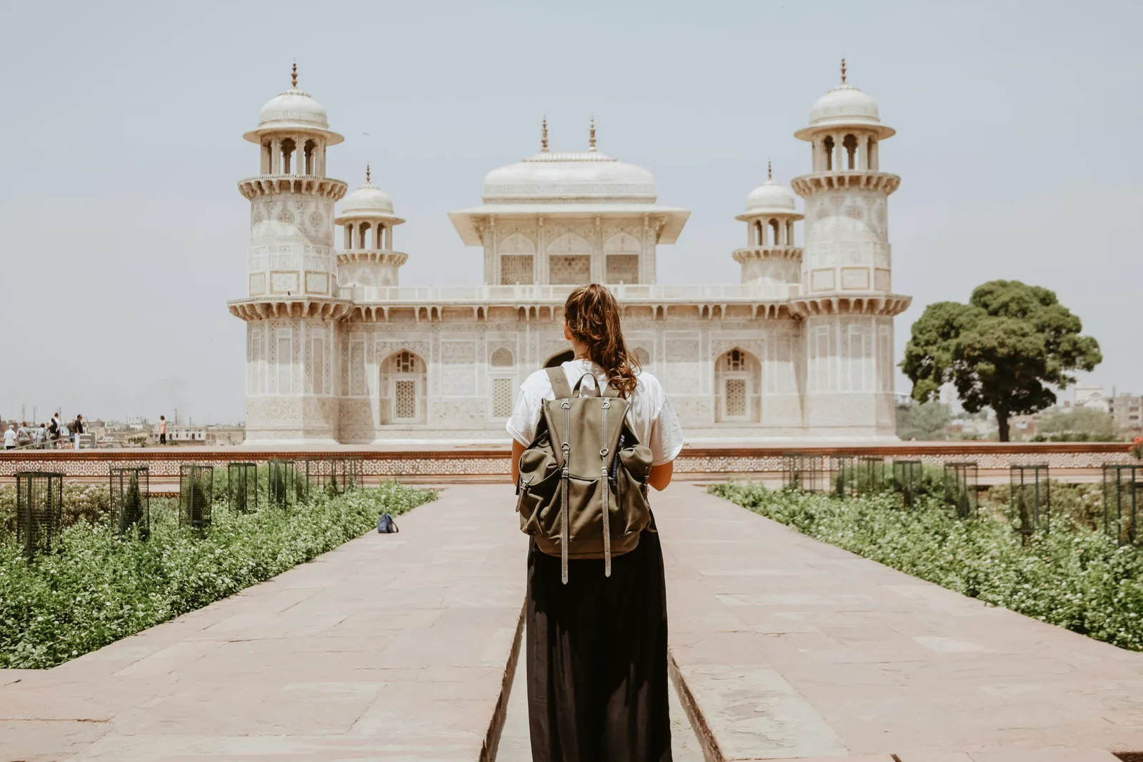 A traveller with a backpack standing on a stone path facing a white marble Mughal tomb.